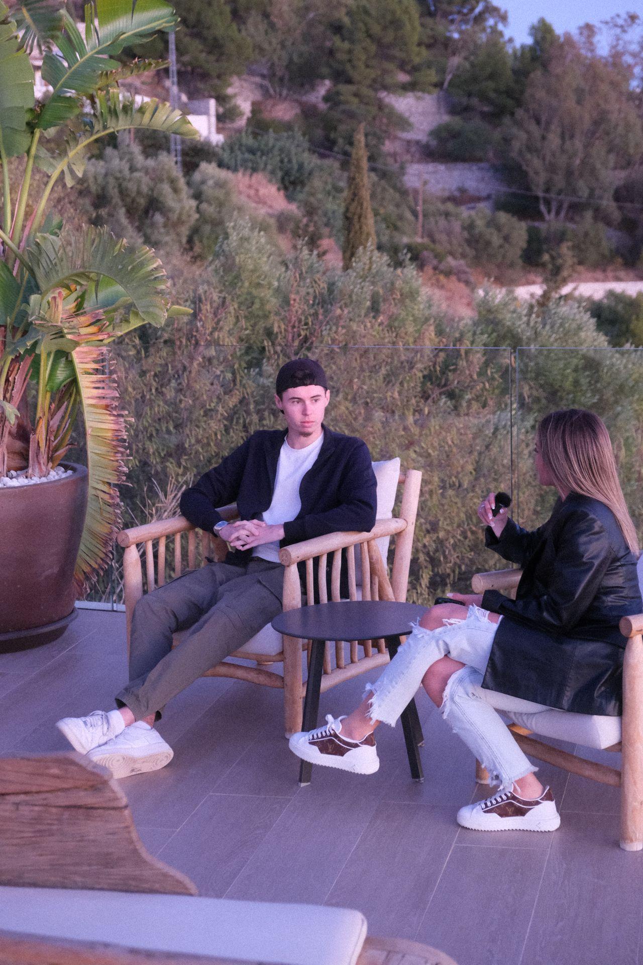 Two people sitting on wooden chairs on a patio surrounded by greenery, having a conversation.